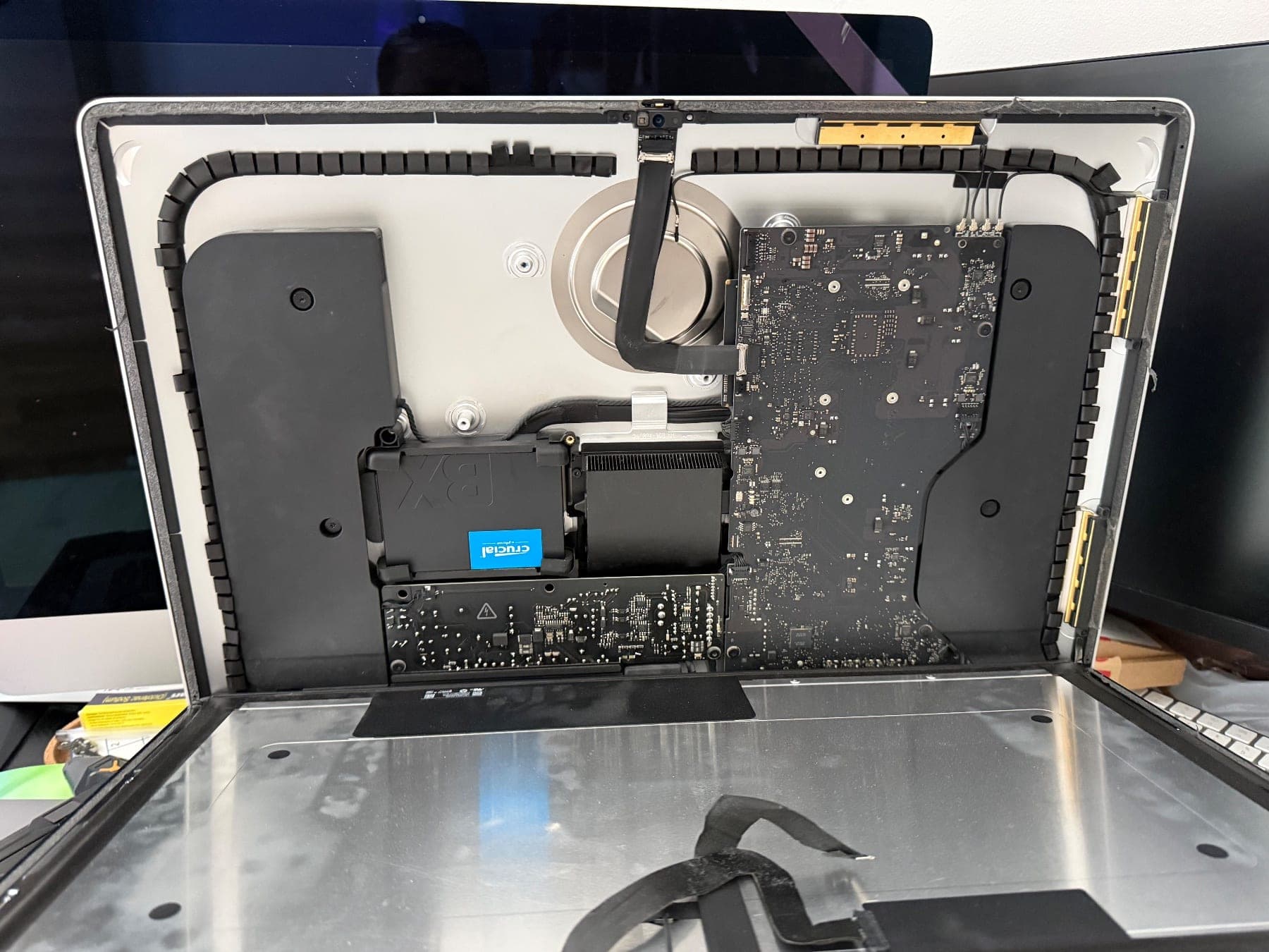 iMac opened with the display panel lifted away, showing the logic board, power supply and a Crucial SSD fitted next to the fan