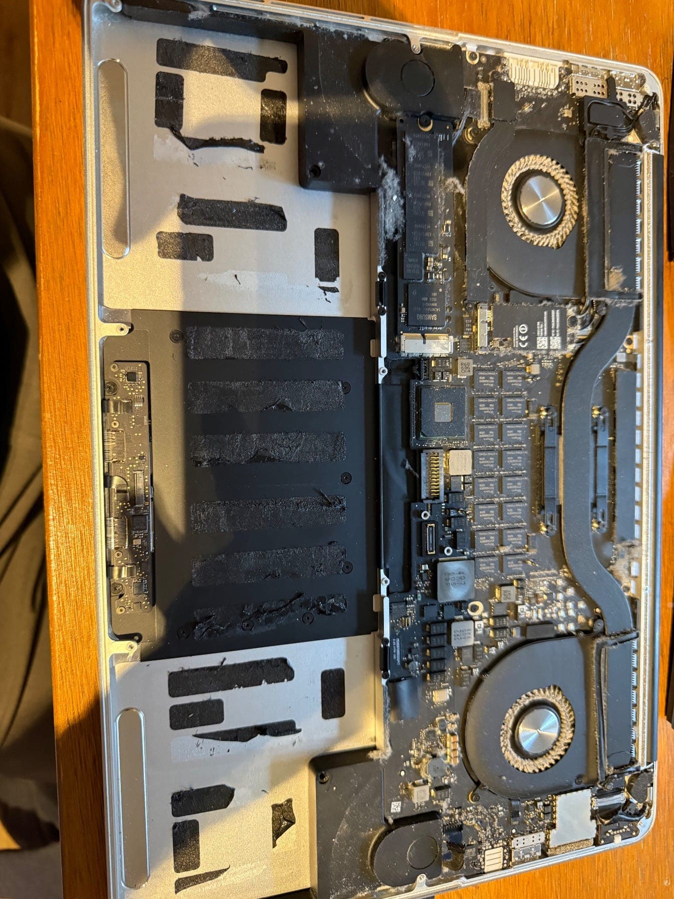 MacBook Pro top case with the battery removed, showing adhesive strips and dust and lint built up around the fans and heatsink