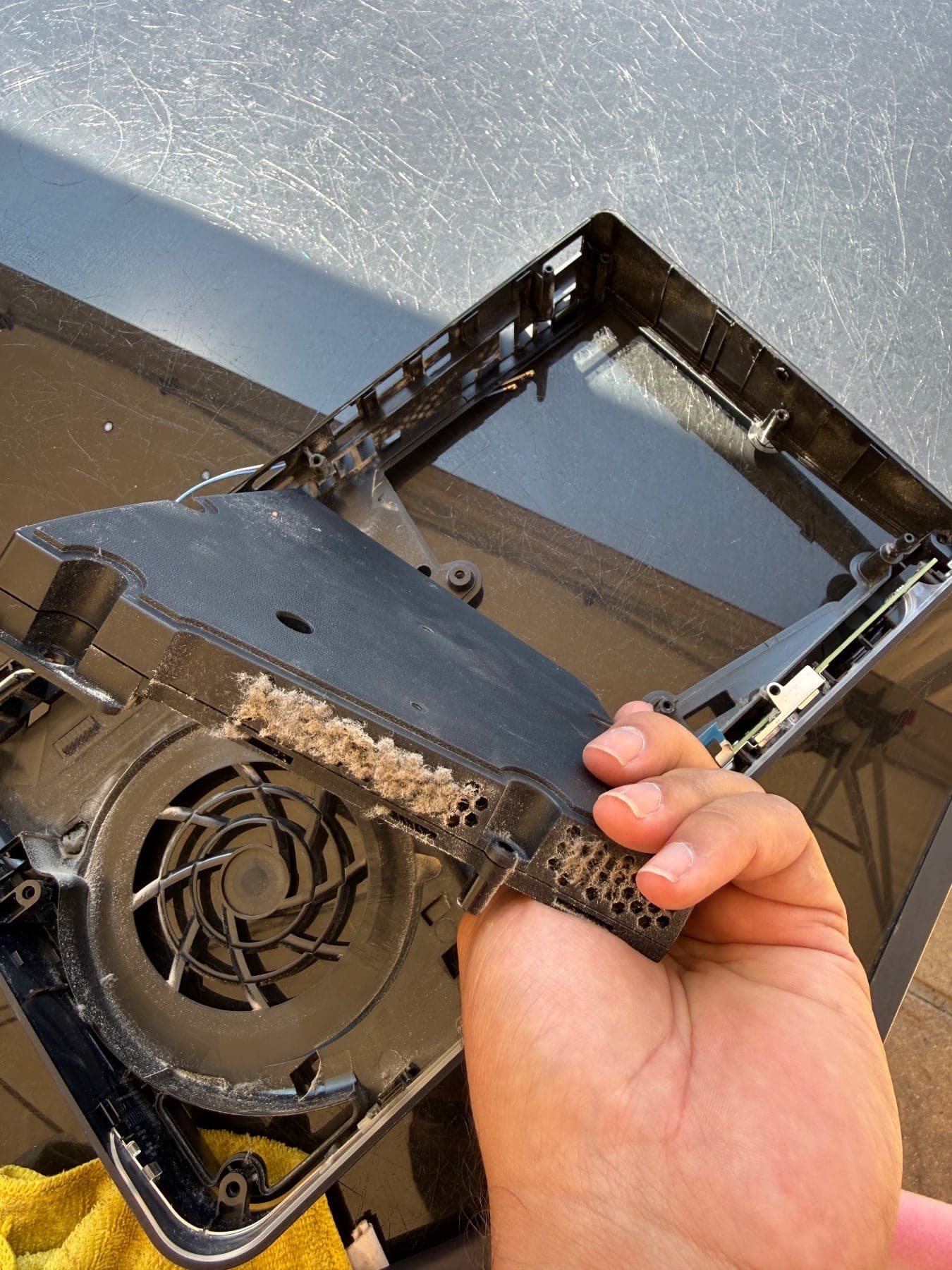 PS5 outer case panel held up showing a line of caked dust along the vent, with the dusty fan visible in the chassis below